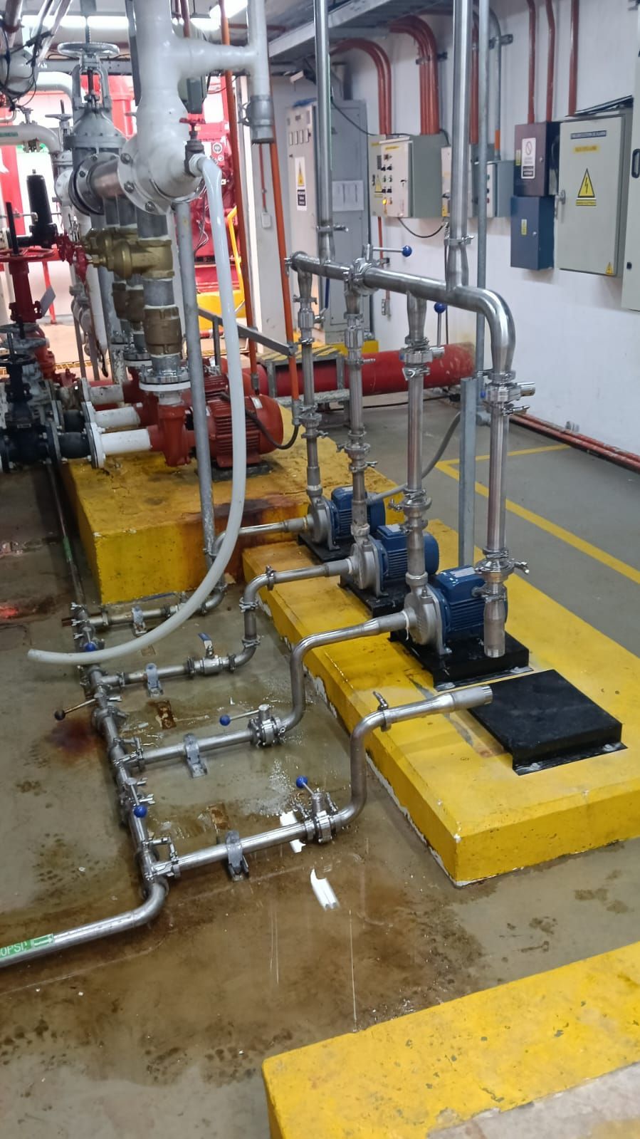 Tuberías y bombas industriales sobre plataformas amarillas en una sala de máquinas húmeda.