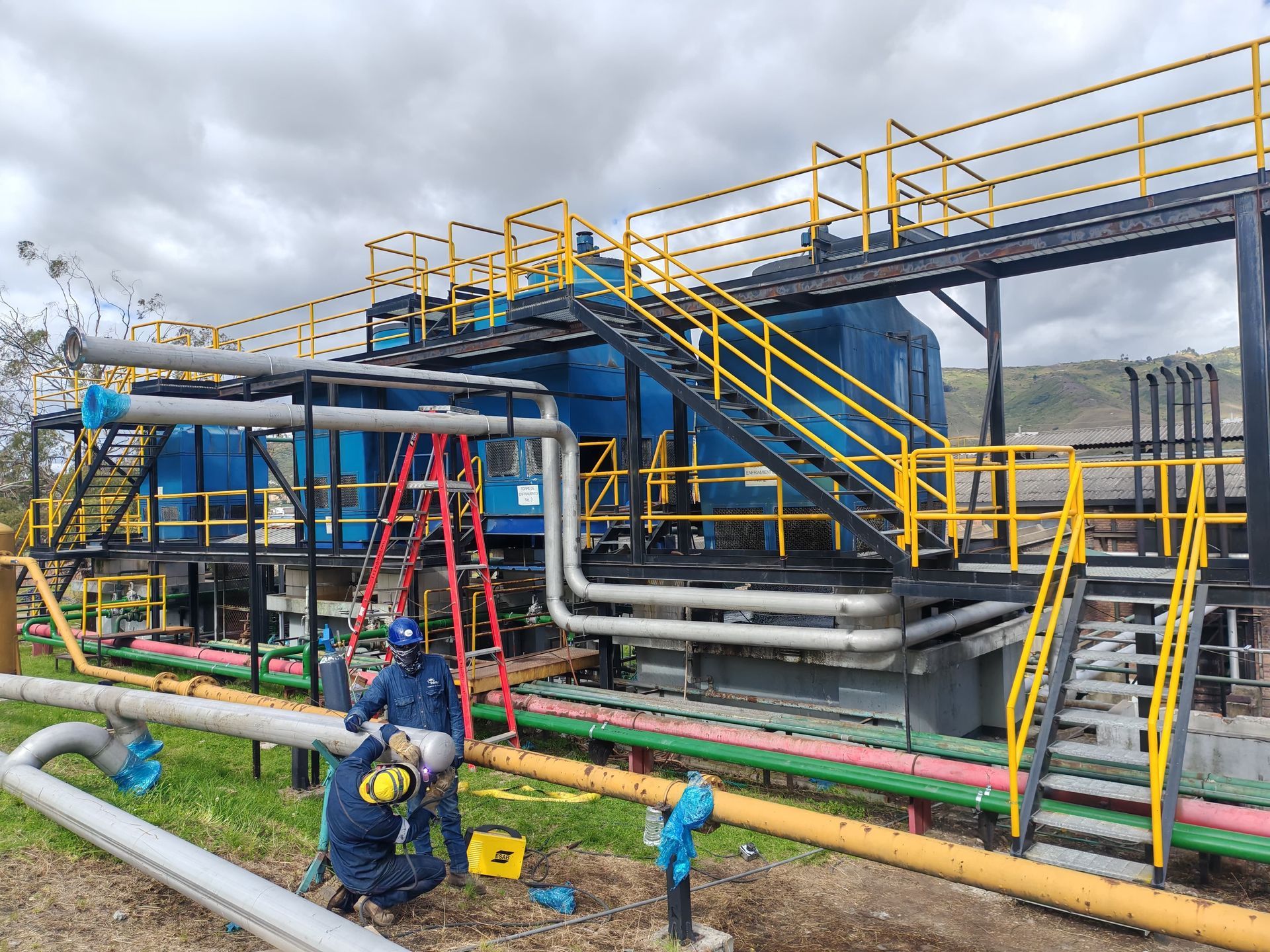 Planta industrial con trabajadores con cascos junto a tuberías y escaleras metálicas azules y amarillas.
