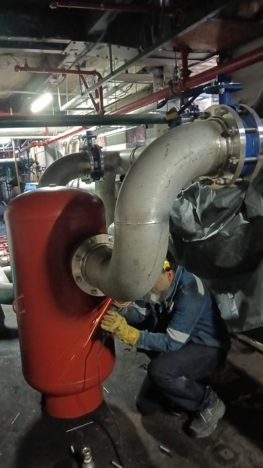 Un trabajador inspecciona con una linterna las tuberías industriales rojas y grises de un cuarto de servicio.