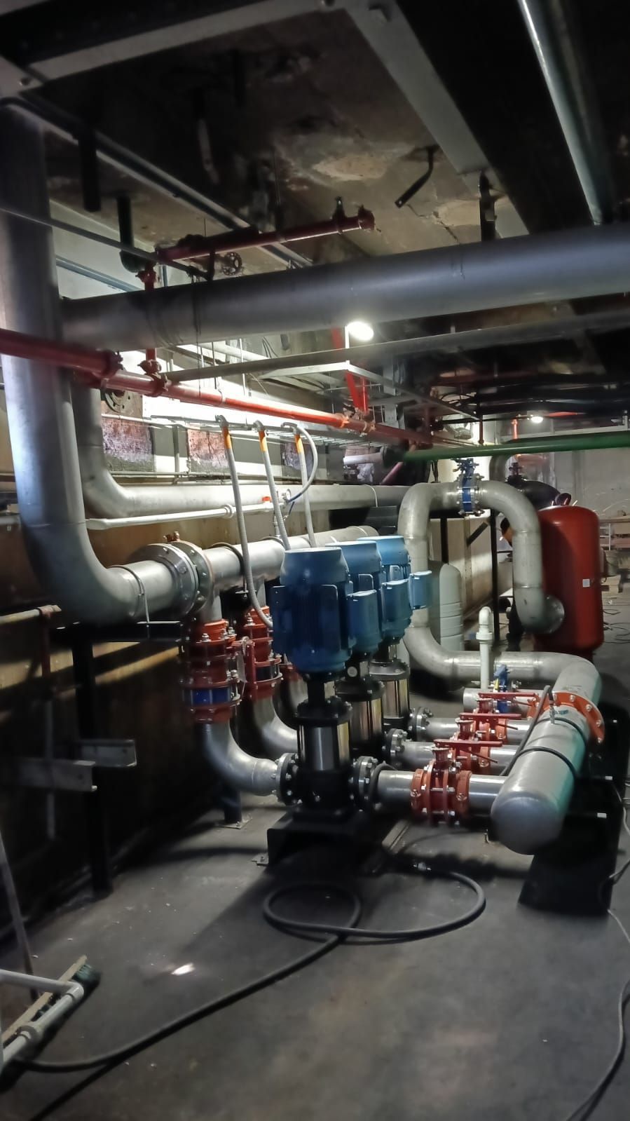 Sala de máquinas en el sótano con grandes tuberías, válvulas, bombas y equipos industriales bajo la tubería superior.