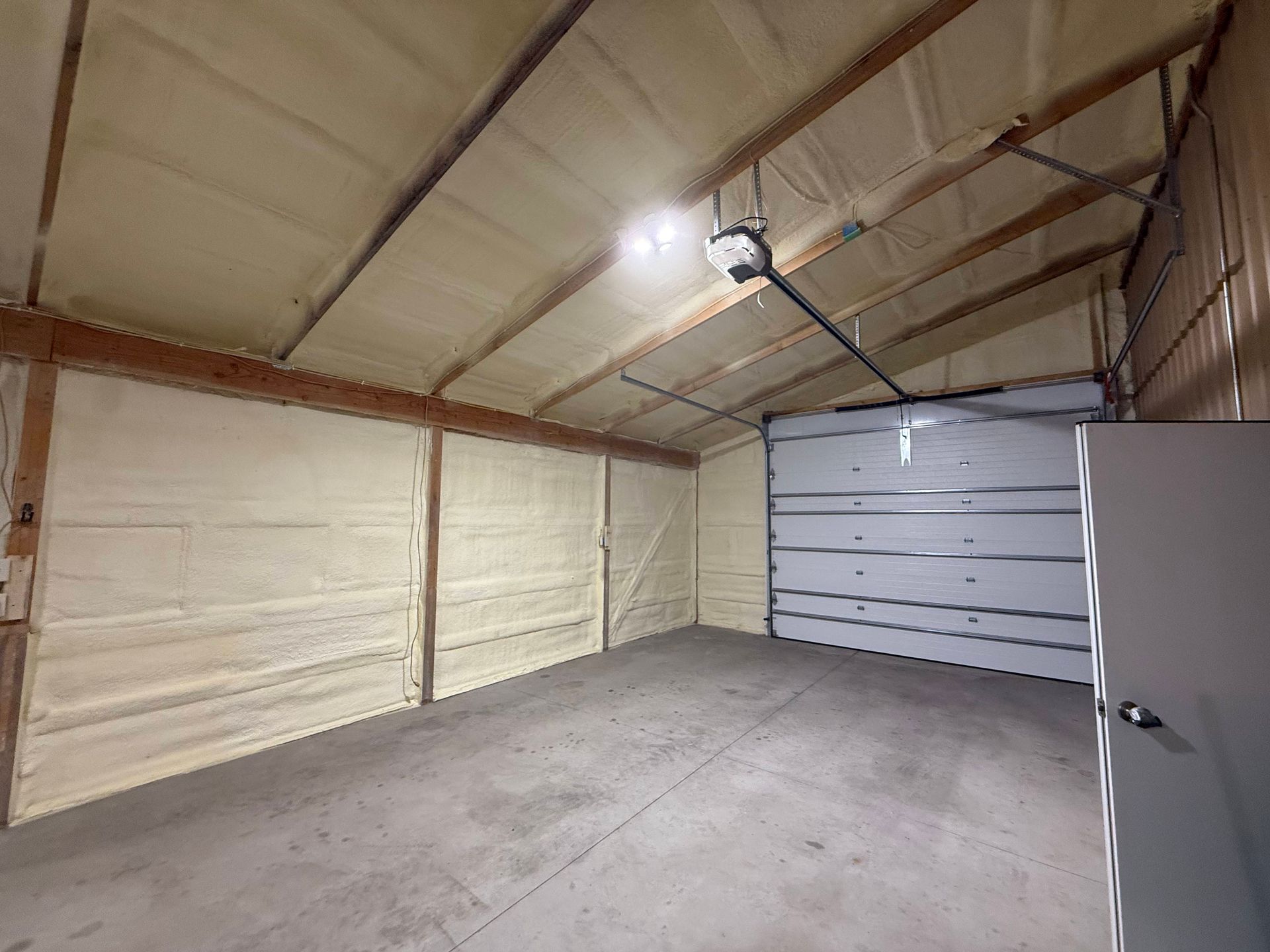 An empty garage with insulated walls and ceiling, concrete flooring, a white roll-up door, and an overhead light.