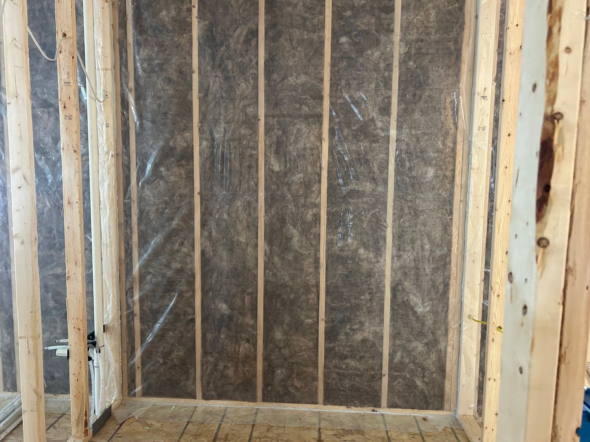 Framed wooden wall studs with brown insulation batts installed between them, partially covered by a thin plastic film.