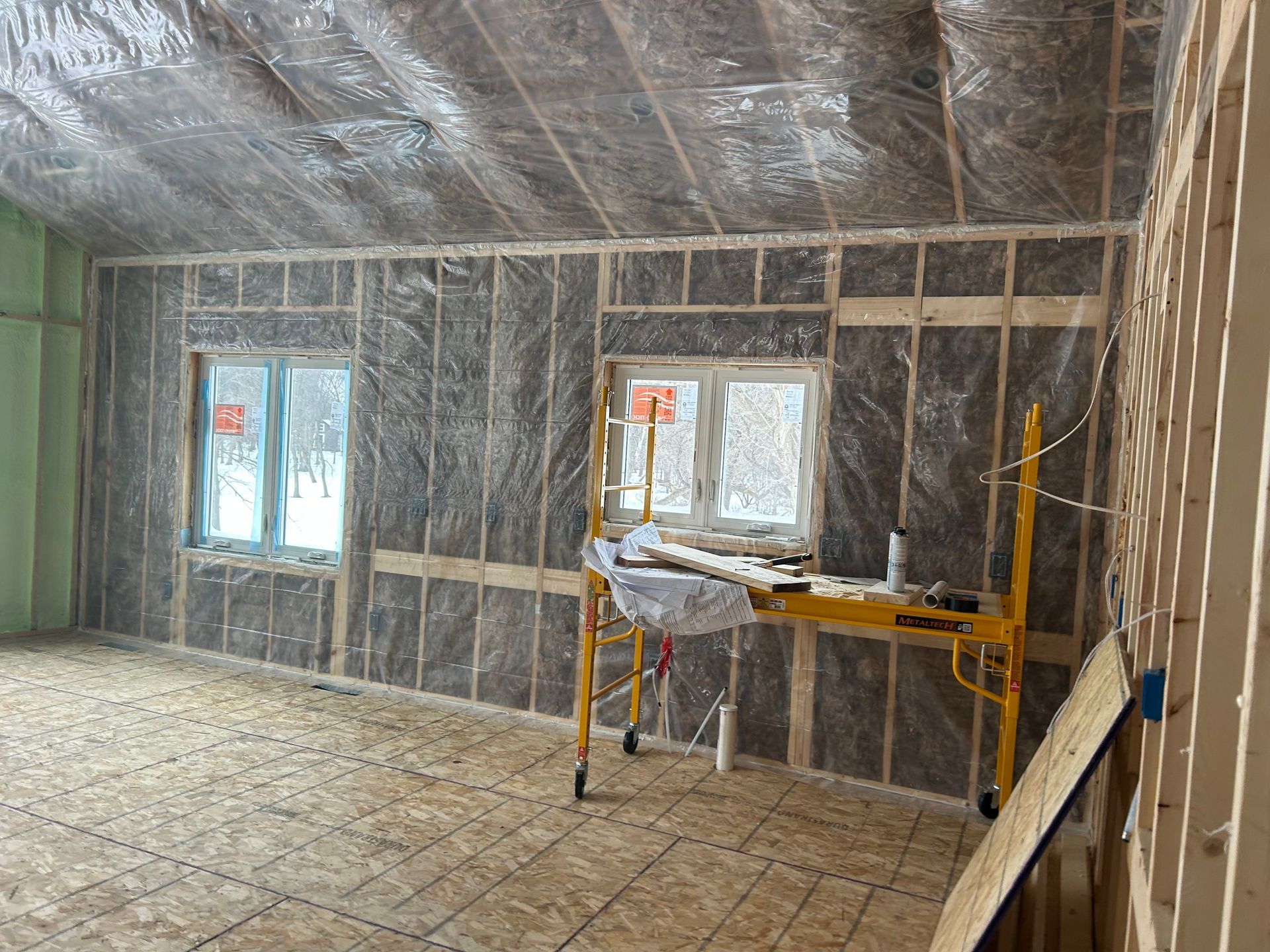 Interior view of a room under construction with insulated walls, wood framing, a subfloor, and yellow scaffolding.