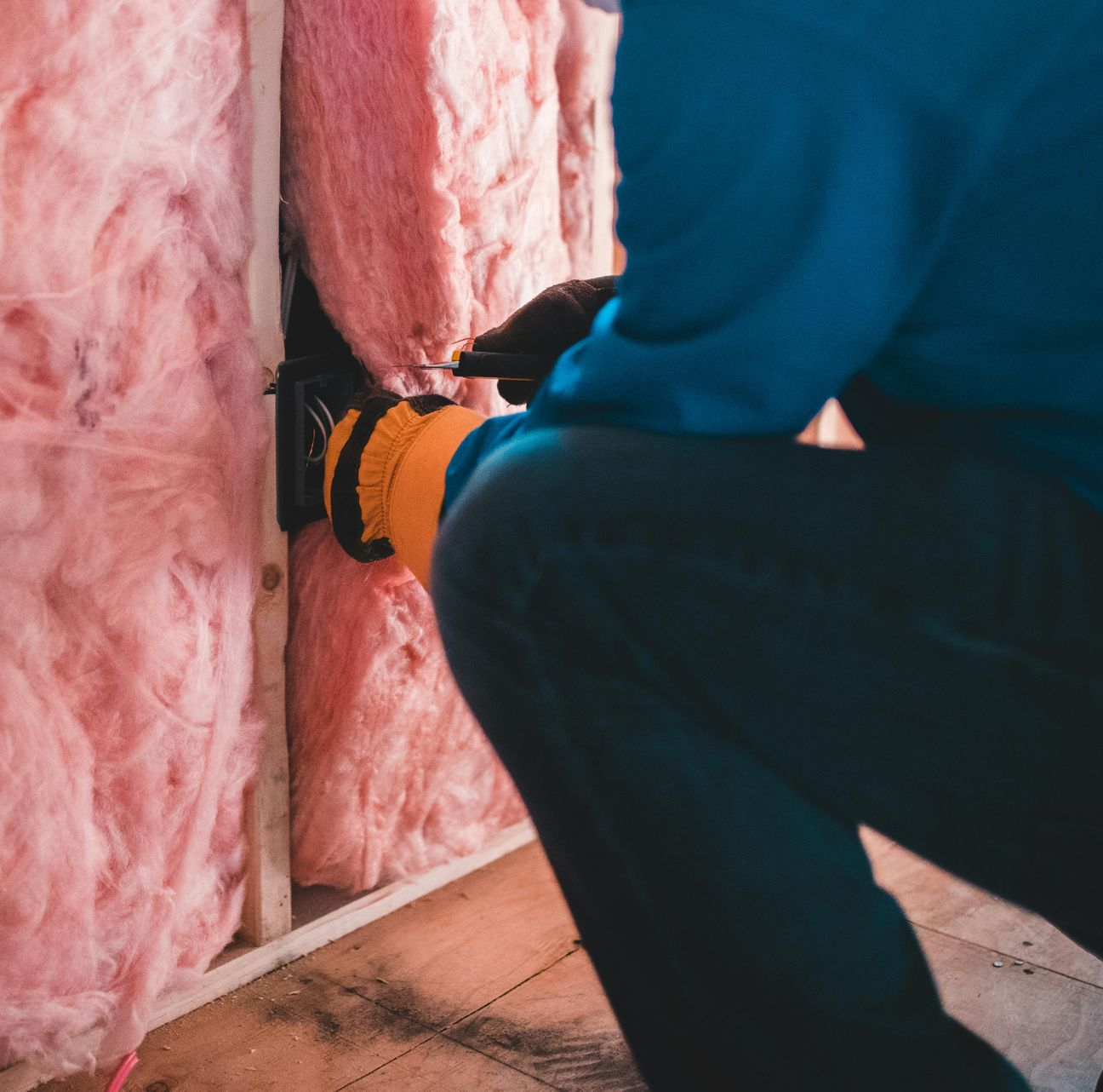 A person in a blue shirt and dark jeans kneels to install pink fiberglass insulation into a wooden wall frame.