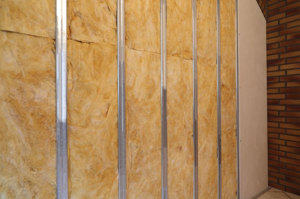 A partial wall assembly featuring metal studs, yellow fiberglass insulation, and one installed drywall panel against brick.