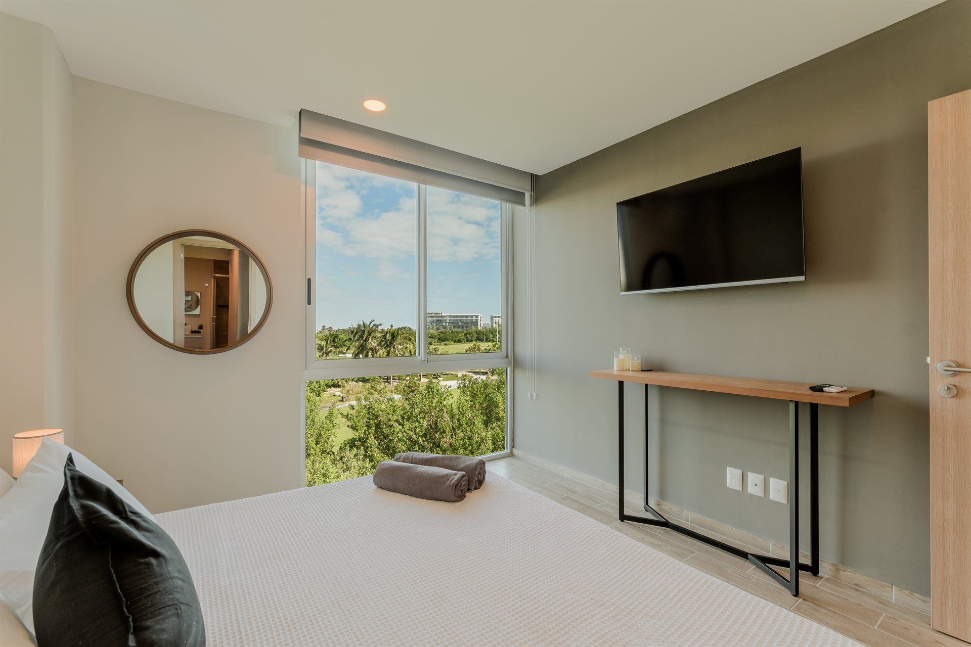 Un dormitorio con cama, televisión, espejo y gran ventanal.