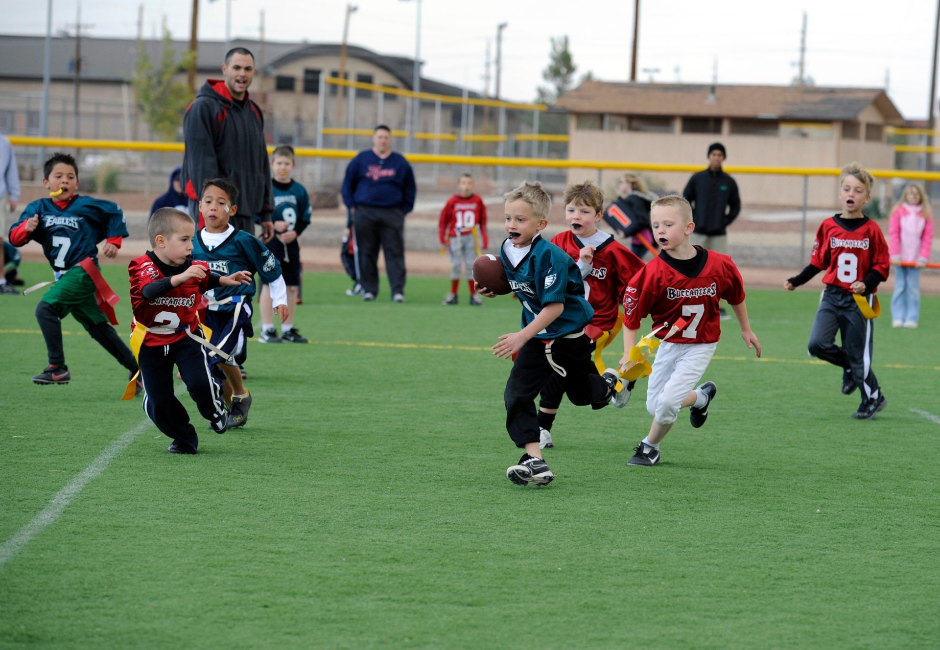 Flag football game at night, a player in red jersey runs with the ball as others pursue on a green field.