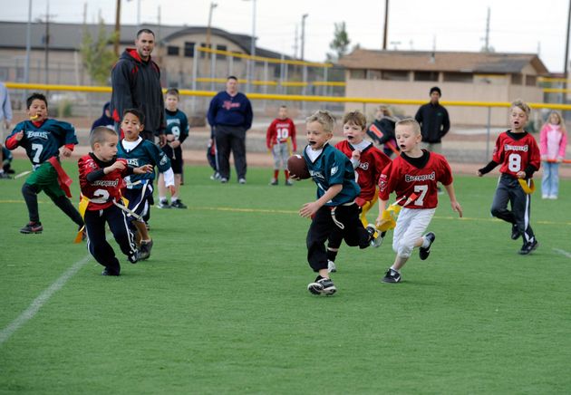 Flag football game at night, a player in red jersey runs with the ball as others pursue on a green field.