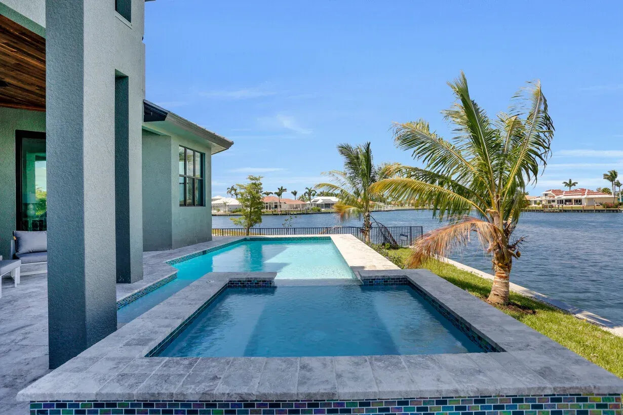Modern waterfront patio with a tiled pool, palm trees, and a house beside the water