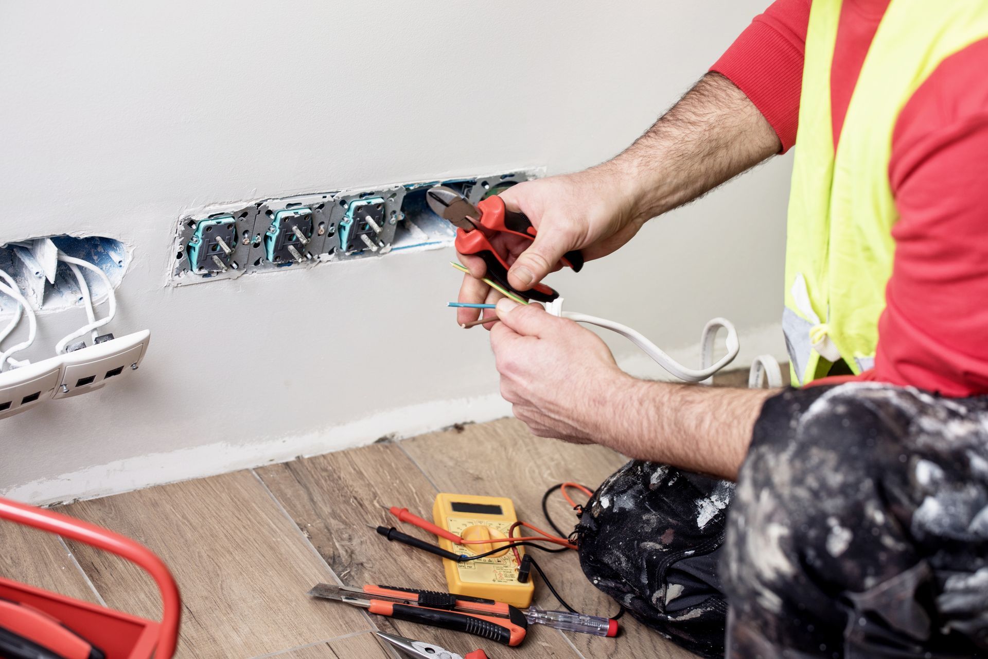 Electrician wiring wall outlet boxes with pliers, multimeter and hand tools nearby.