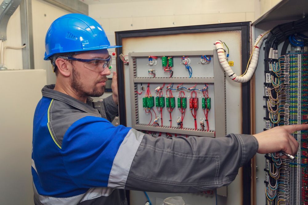 Electrician in blue hard hat examining an electrical panel, pointing at wires.
