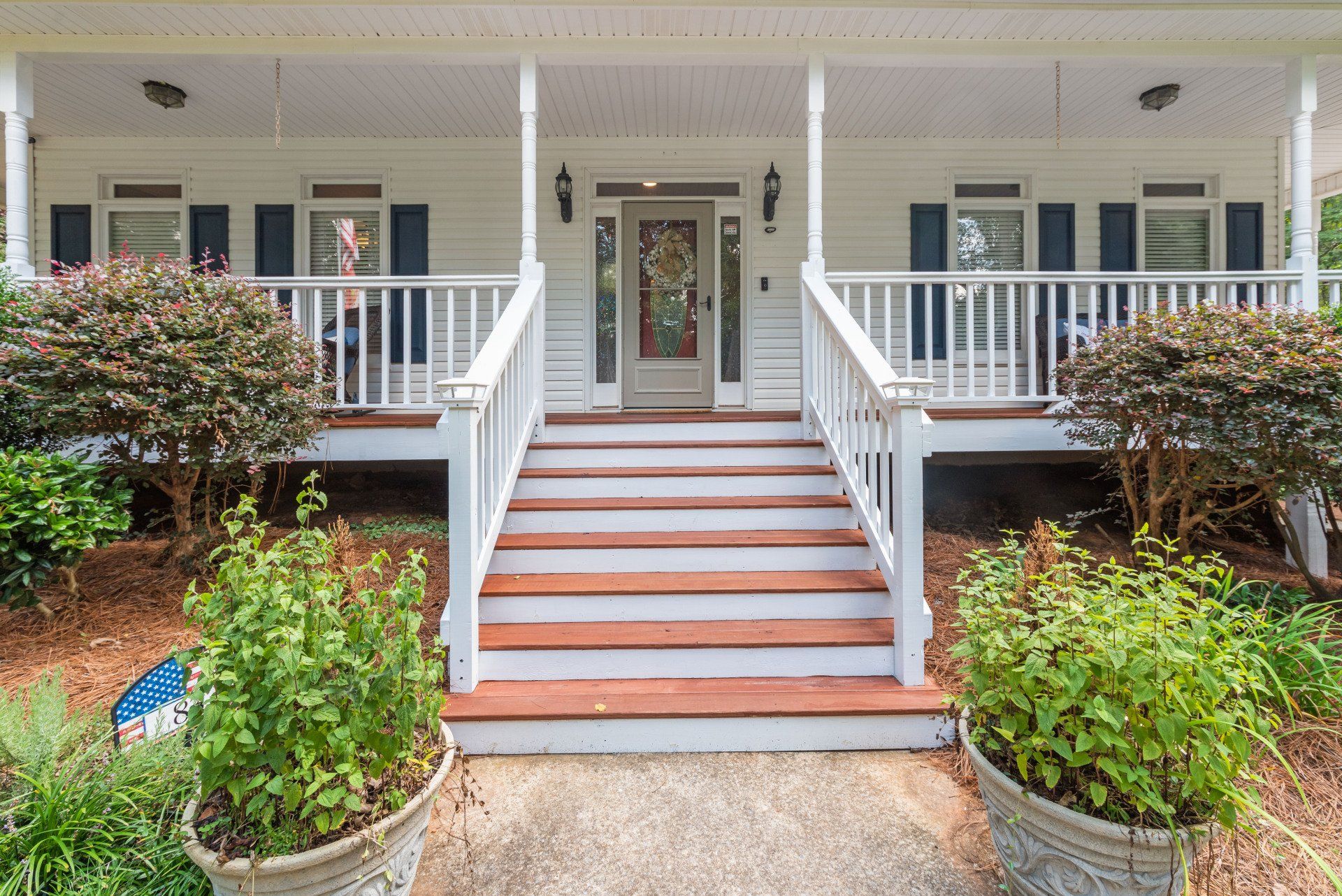 White house with front porch, steps, and door. Bushes and planters surround the entrance.