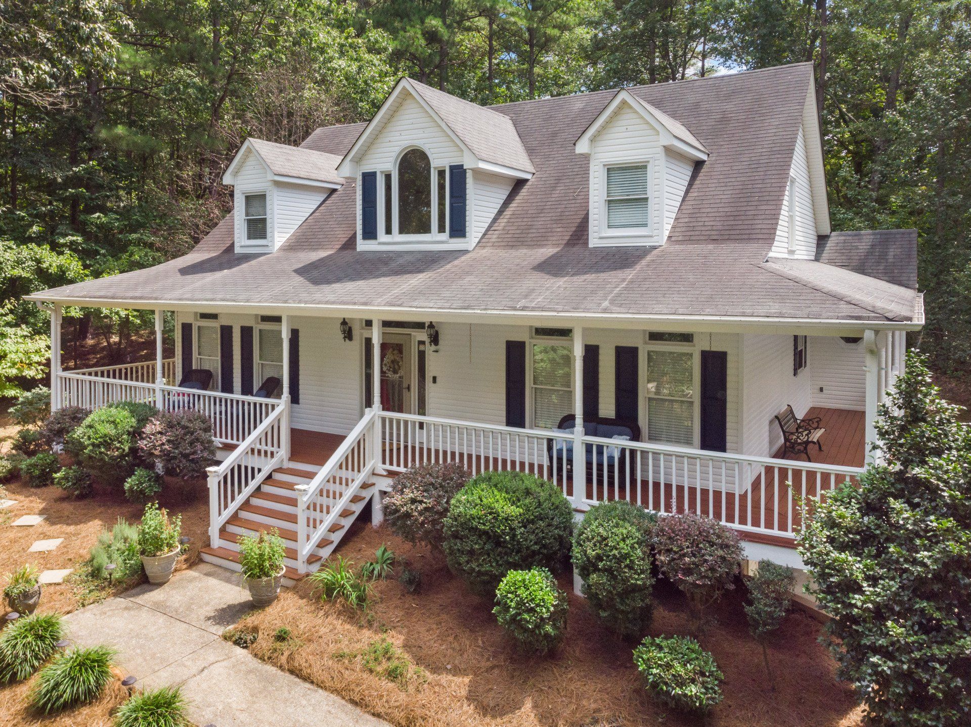 White house with wrap-around porch, shutters, and dormers. Front yard with plants, trees.