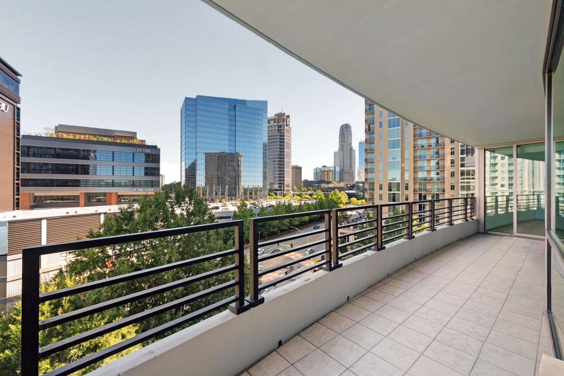 Balcony overlooking a city skyline. Buildings, trees, and blue sky visible. Black railing and tiled floor.
