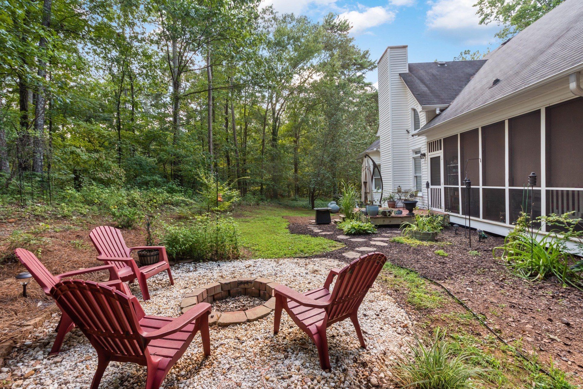 Backyard with red Adirondack chairs around a fire pit, next to a house with a screened porch, surrounded by trees.