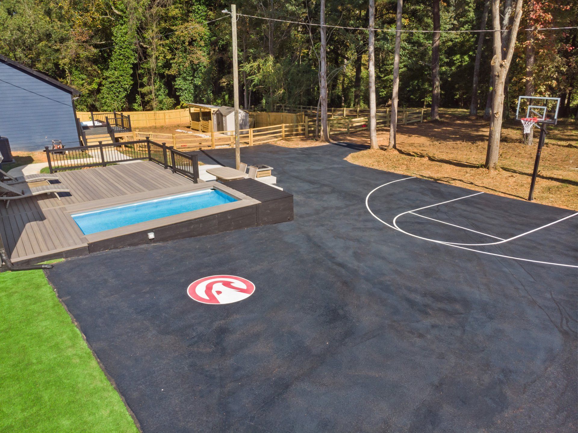 Asphalt basketball court with a logo, a small pool, and a deck. Trees in the background.