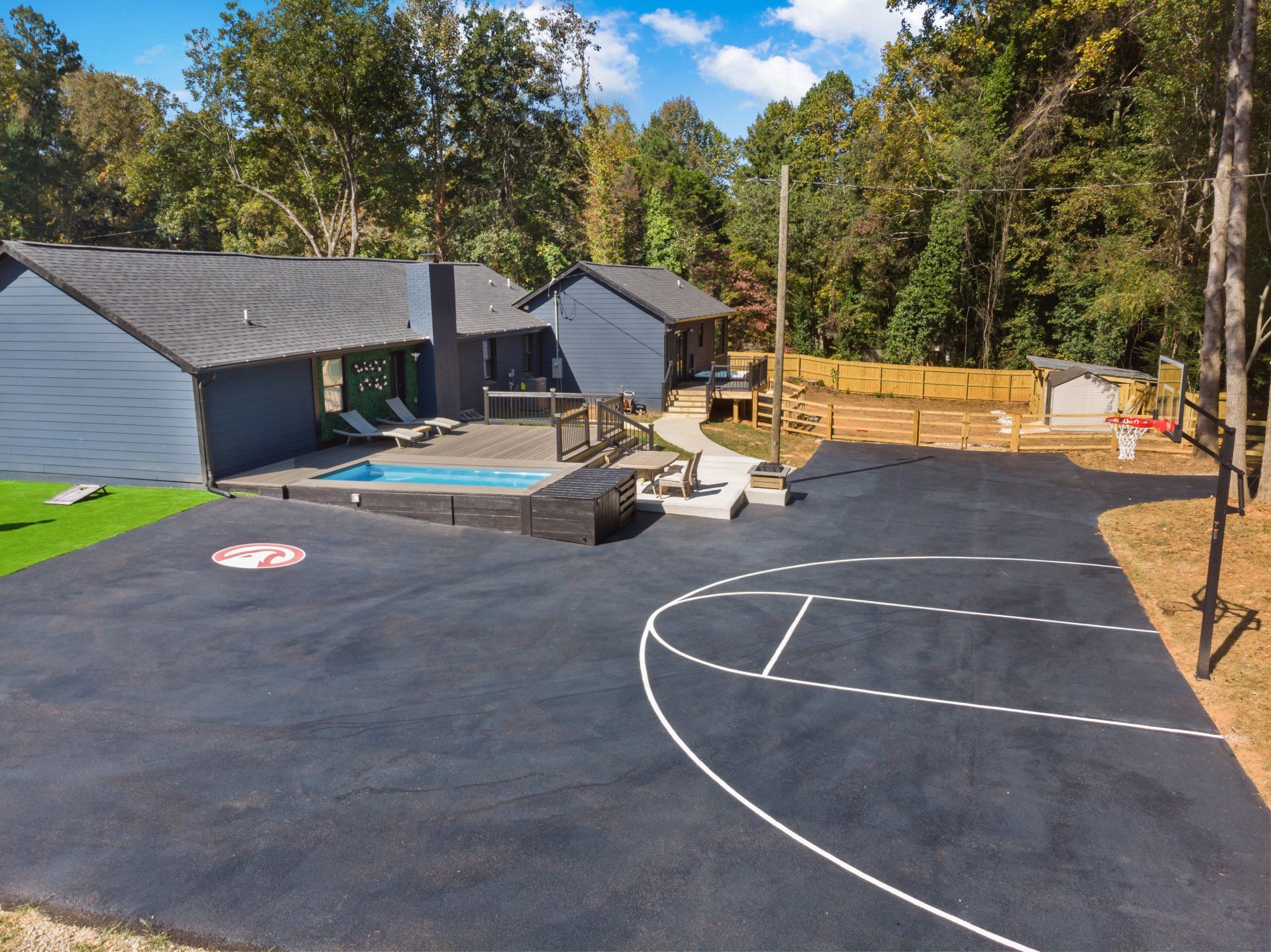 Asphalt basketball court next to a house with a pool in a wooded area.