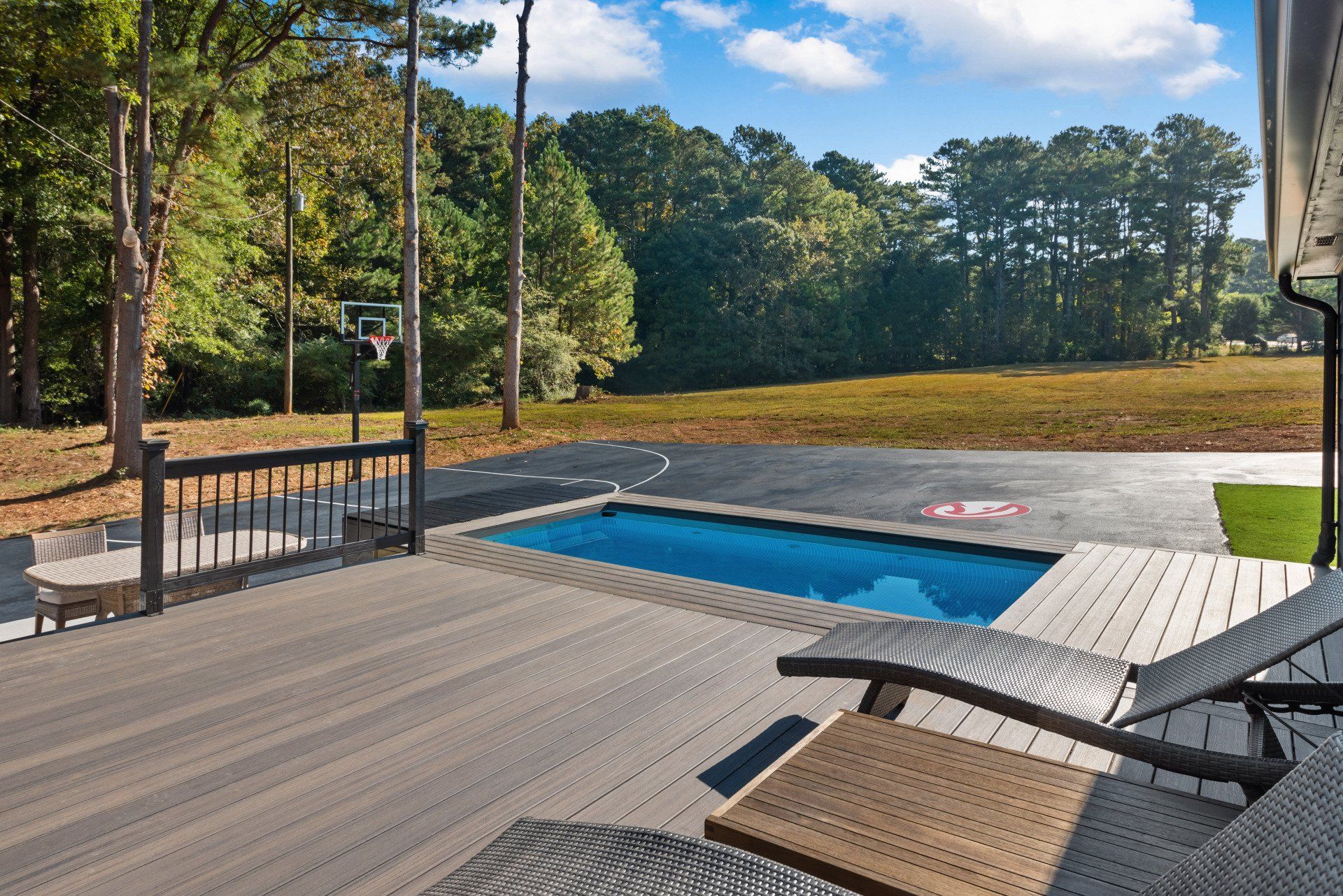 A deck with lounge chairs, a small pool, and a basketball court overlooking a wooded area on a sunny day.