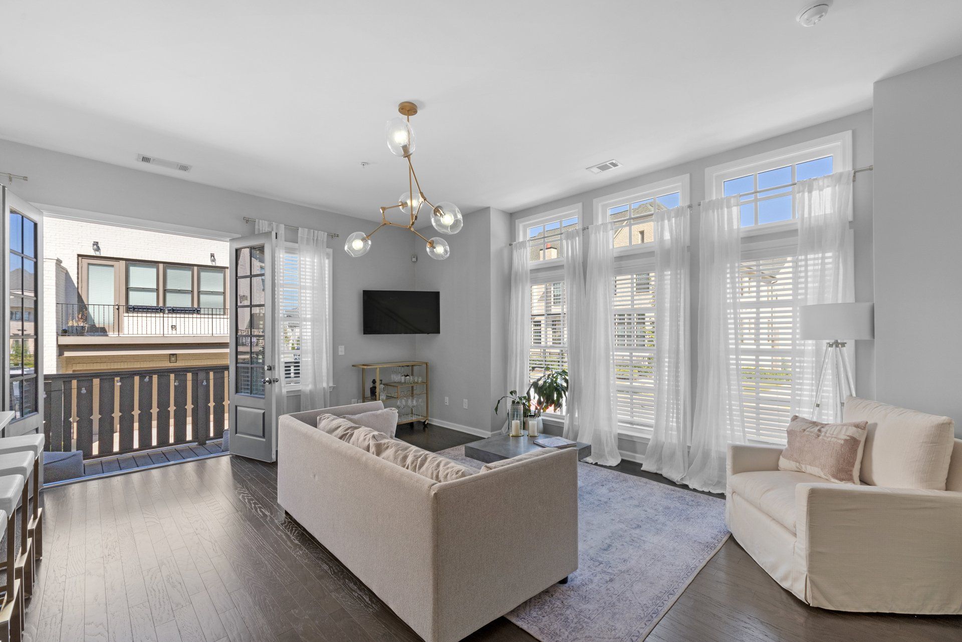 Living room with balcony access, light gray walls, dark wood floors, large windows with sheer curtains.