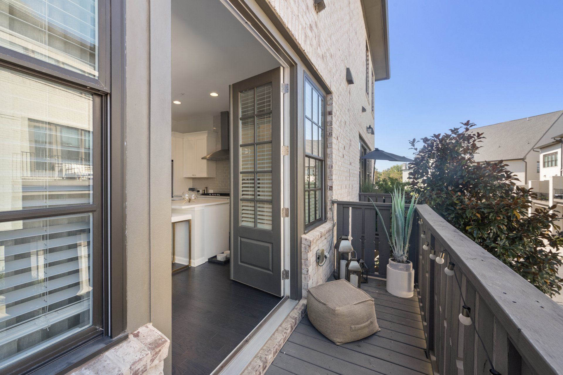 Balcony with dark flooring, open door to kitchen, gray and white exterior. Gray wooden railing with potted plant.