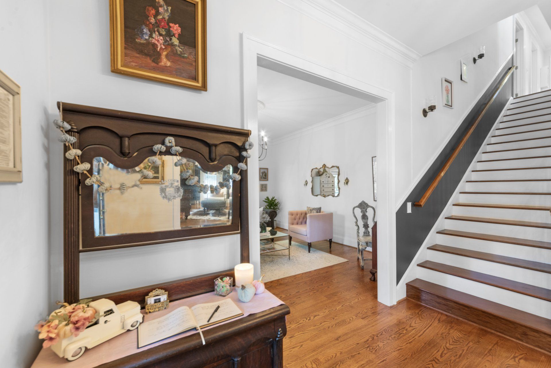Entryway with wooden staircase, mirror, and decorative furniture; view into a living room.