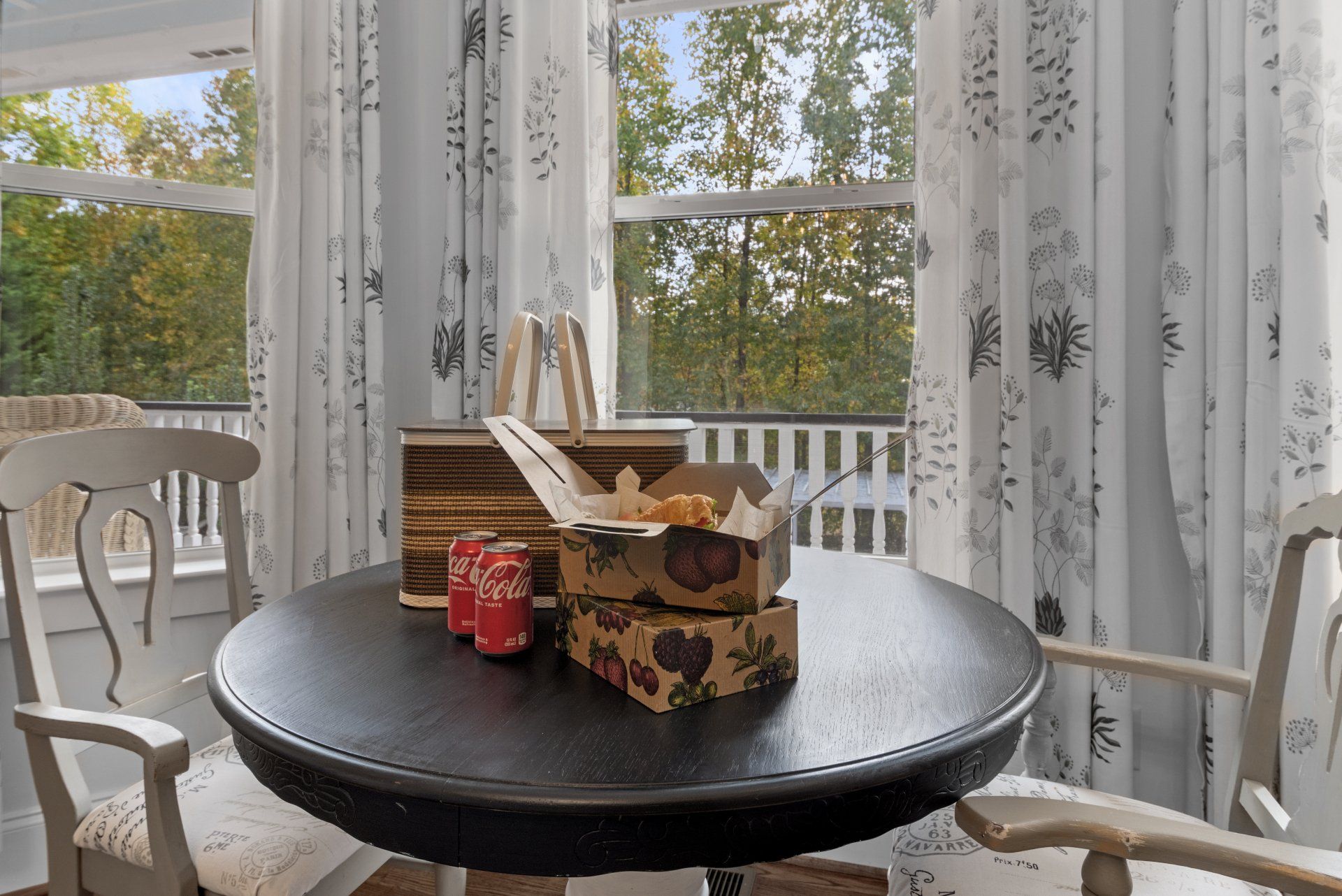 Round table with food boxes and drinks by a window with white curtains, surrounded by chairs.