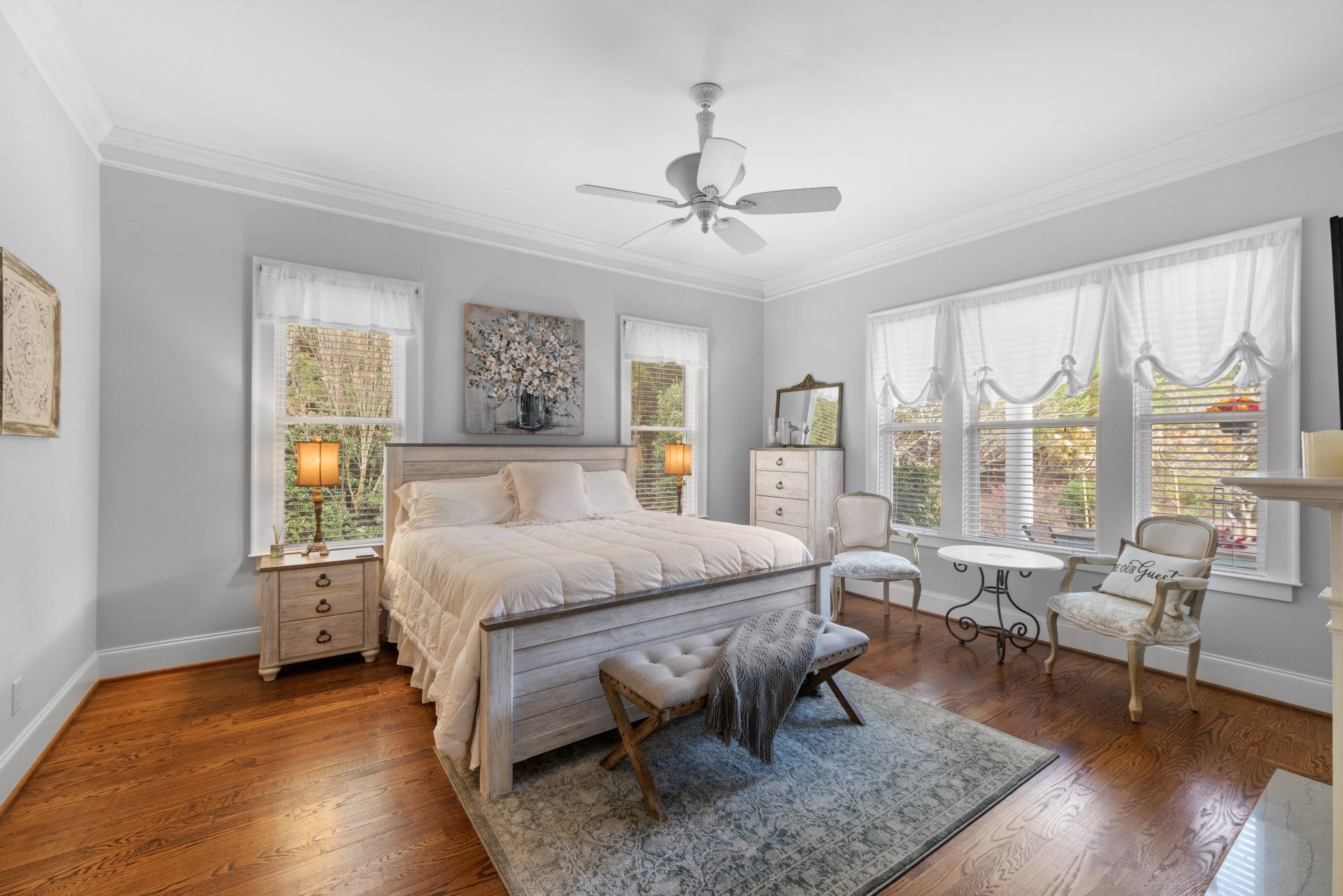 Bedroom with light blue walls, white bed, windows, rug, hardwood floor, and a ceiling fan.