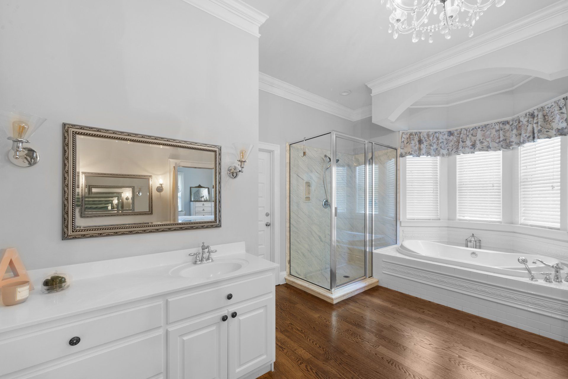 Spacious white bathroom with a vanity, shower, tub, and chandelier. Wooden floor, pale gray walls, and natural light.
