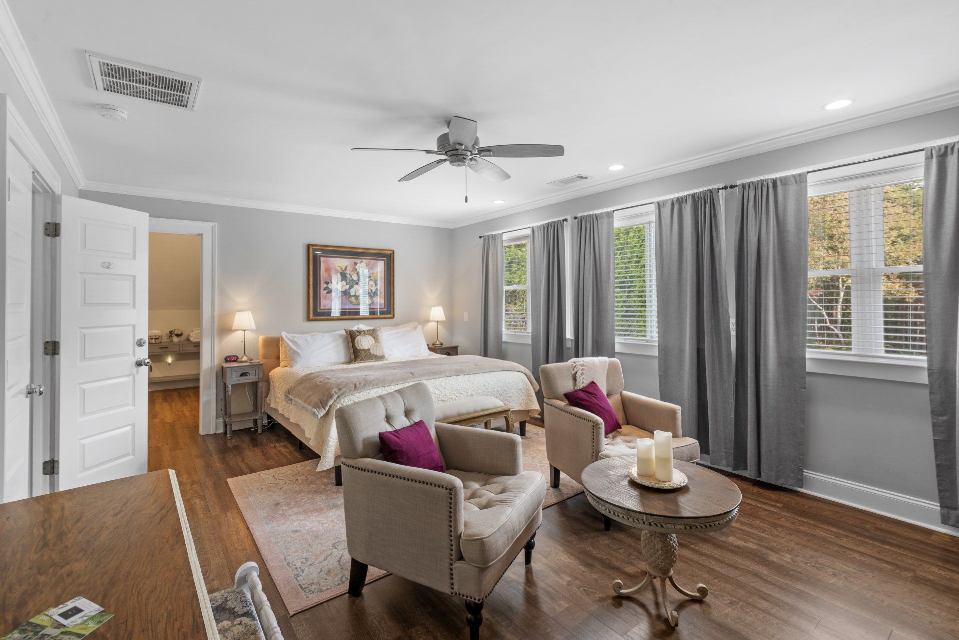 Elegant bedroom with a king bed, sitting area, and large windows. Gray walls, hardwood floors, and gray curtains.