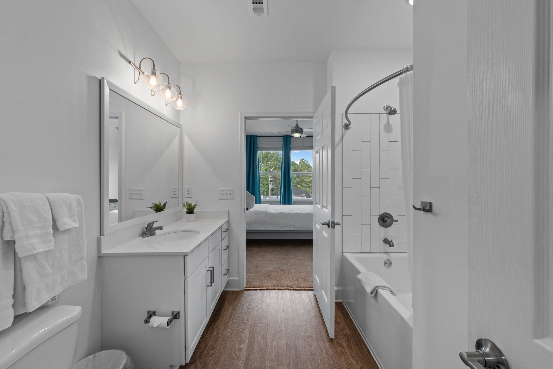 White bathroom with vanity, toilet, and bathtub; door opens to a bedroom.