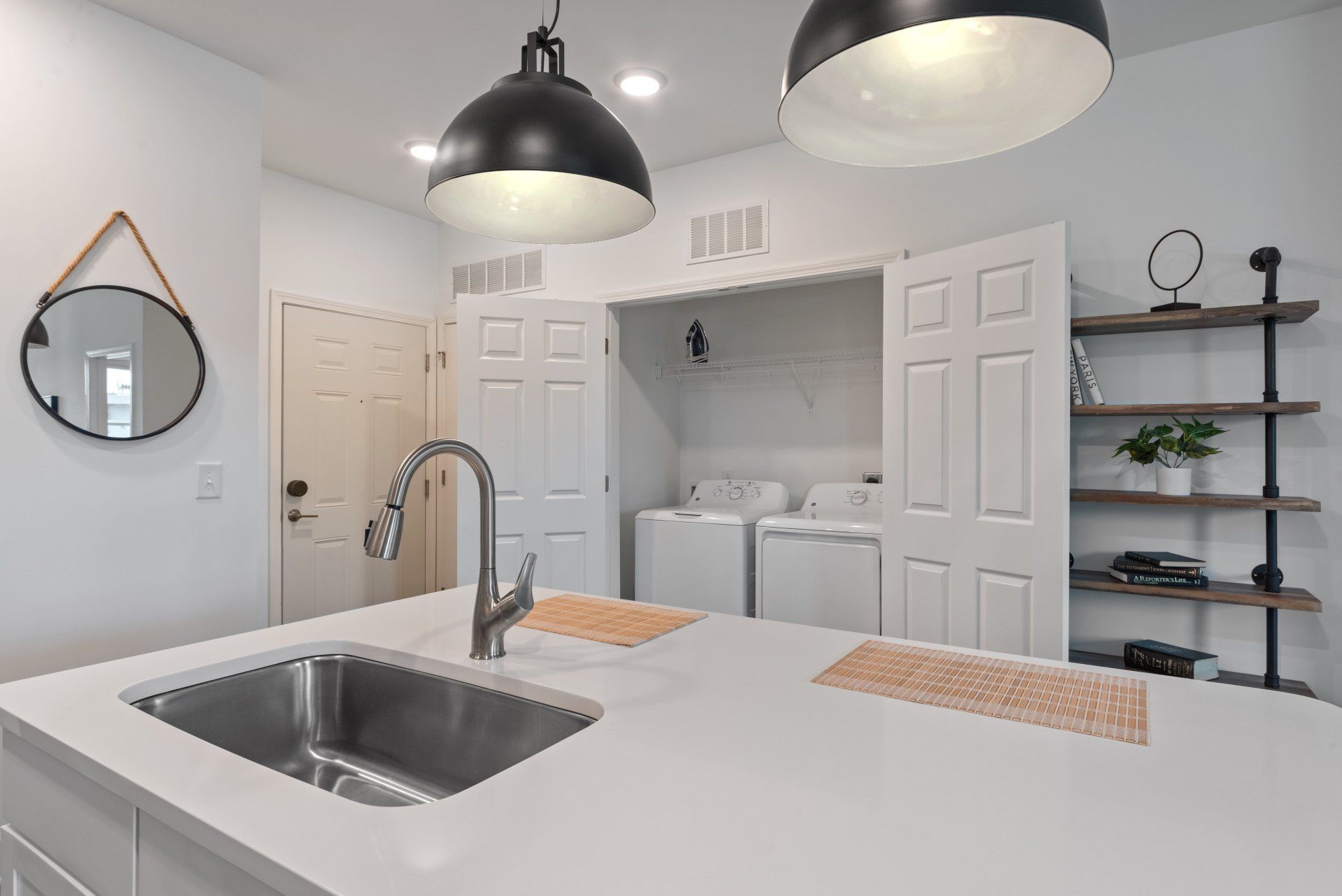 Modern kitchen with a sink, white countertops, black pendant lights, and washer/dryer behind closed doors.