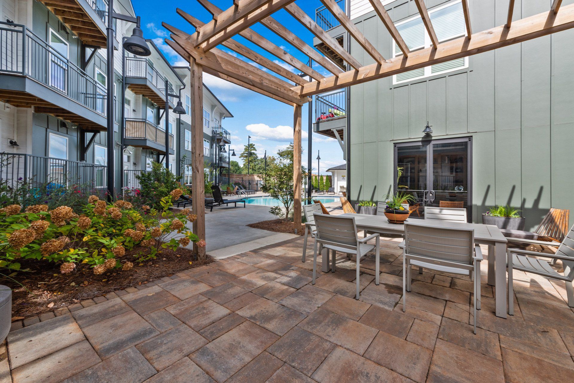 Patio with table and chairs under a pergola, leading to a pool area and buildings.