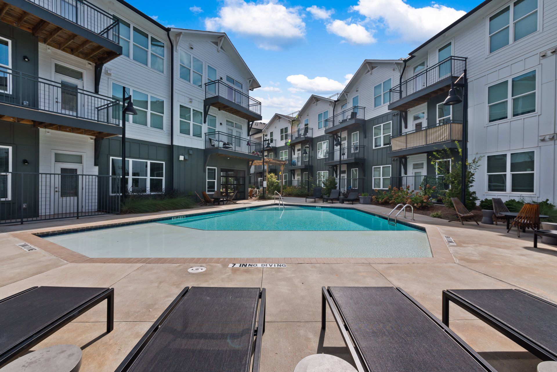 Apartment complex with a pool; lounge chairs in foreground, buildings in background, blue sky.