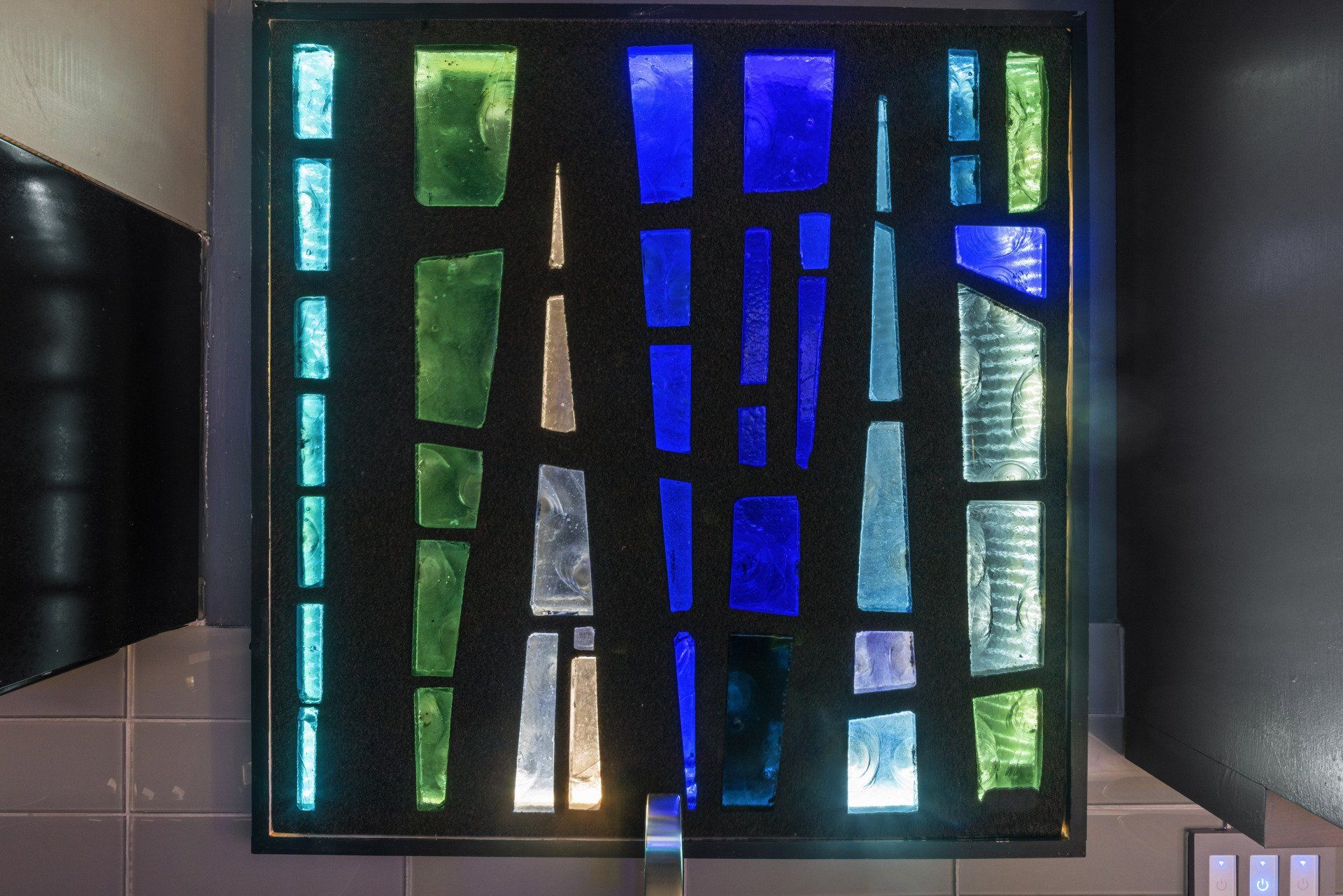 Stained glass artwork with blue, green, and silver rectangles arranged on a black background.