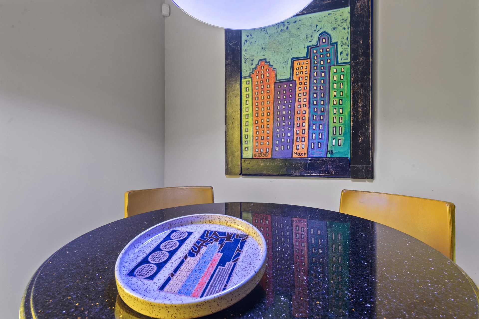Round black table with decorative plate, two chairs, and cityscape art on the wall.