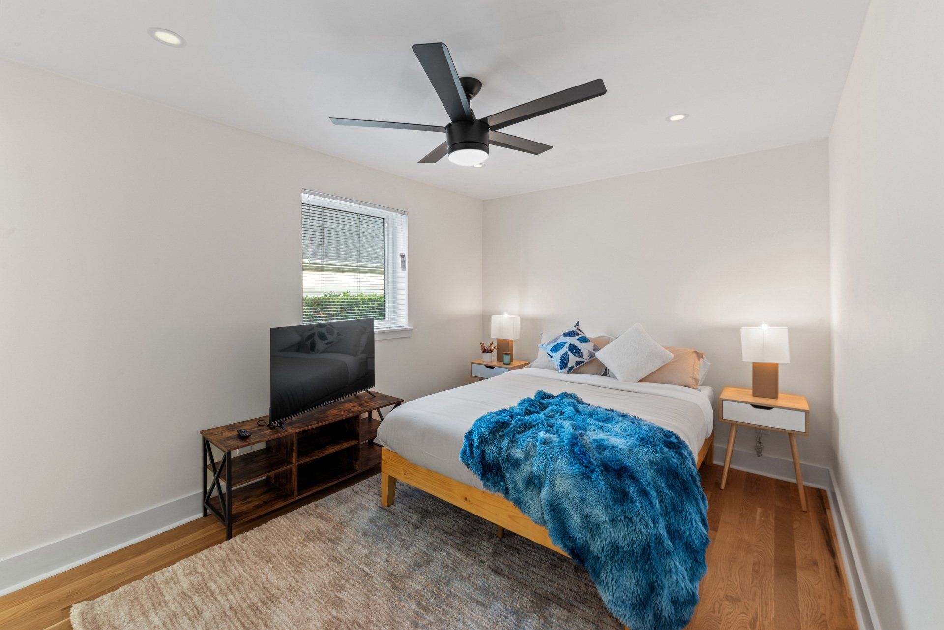 Bedroom with bed, tv, rug, and ceiling fan. White walls, wooden floor, and blue accents.