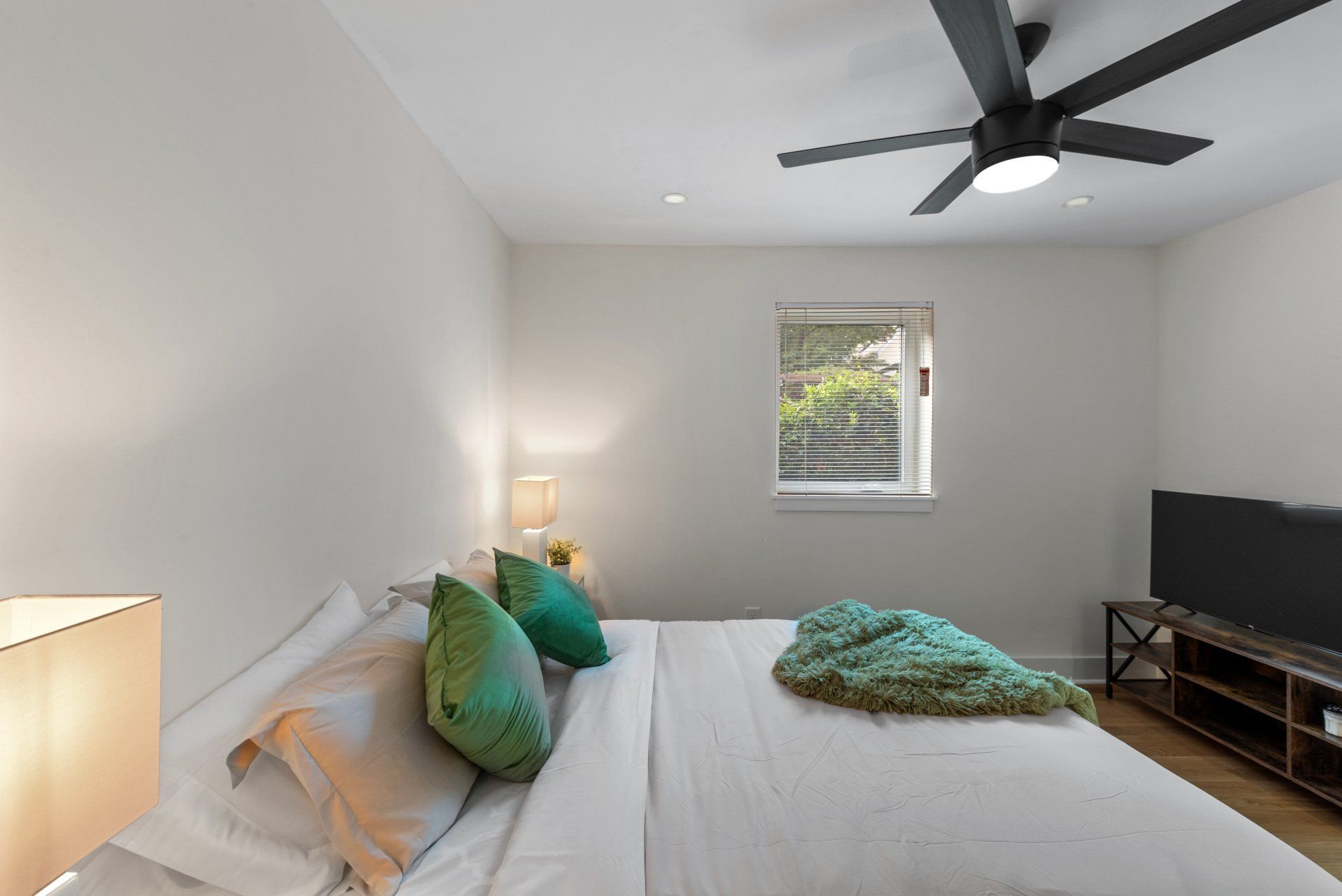Bedroom with a bed, TV, and ceiling fan. White walls, green pillows, and a window.