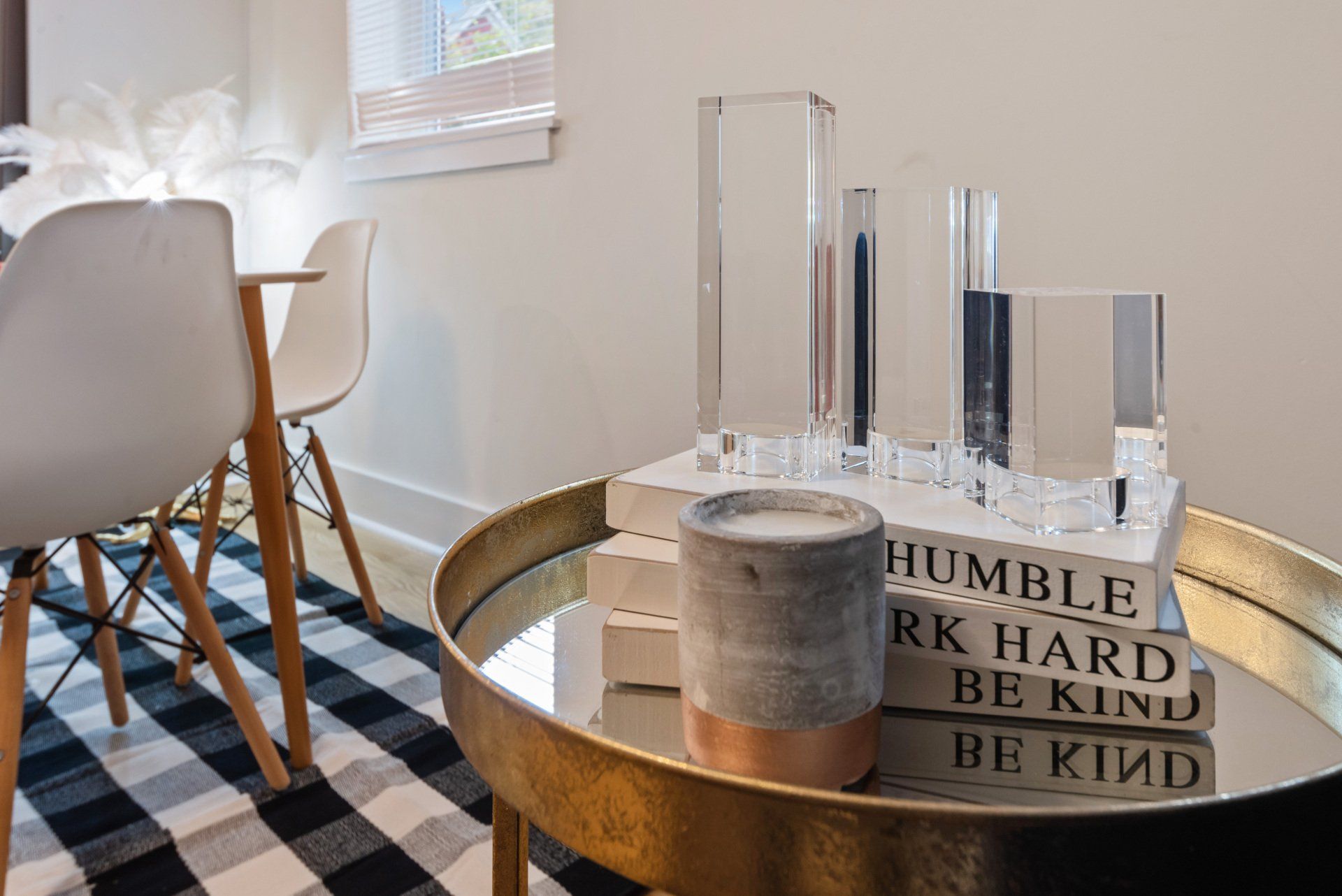 Modern living room scene with decorative table, chairs, and checkered rug. Gold table with clear glass objects and a candle.