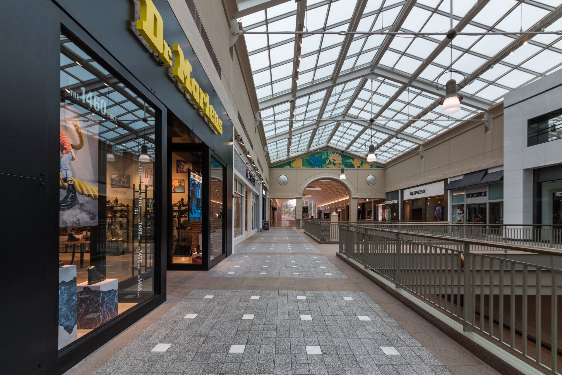 Interior view of a shopping mall hallway with stores on the left and right sides. Glass roof overhead.