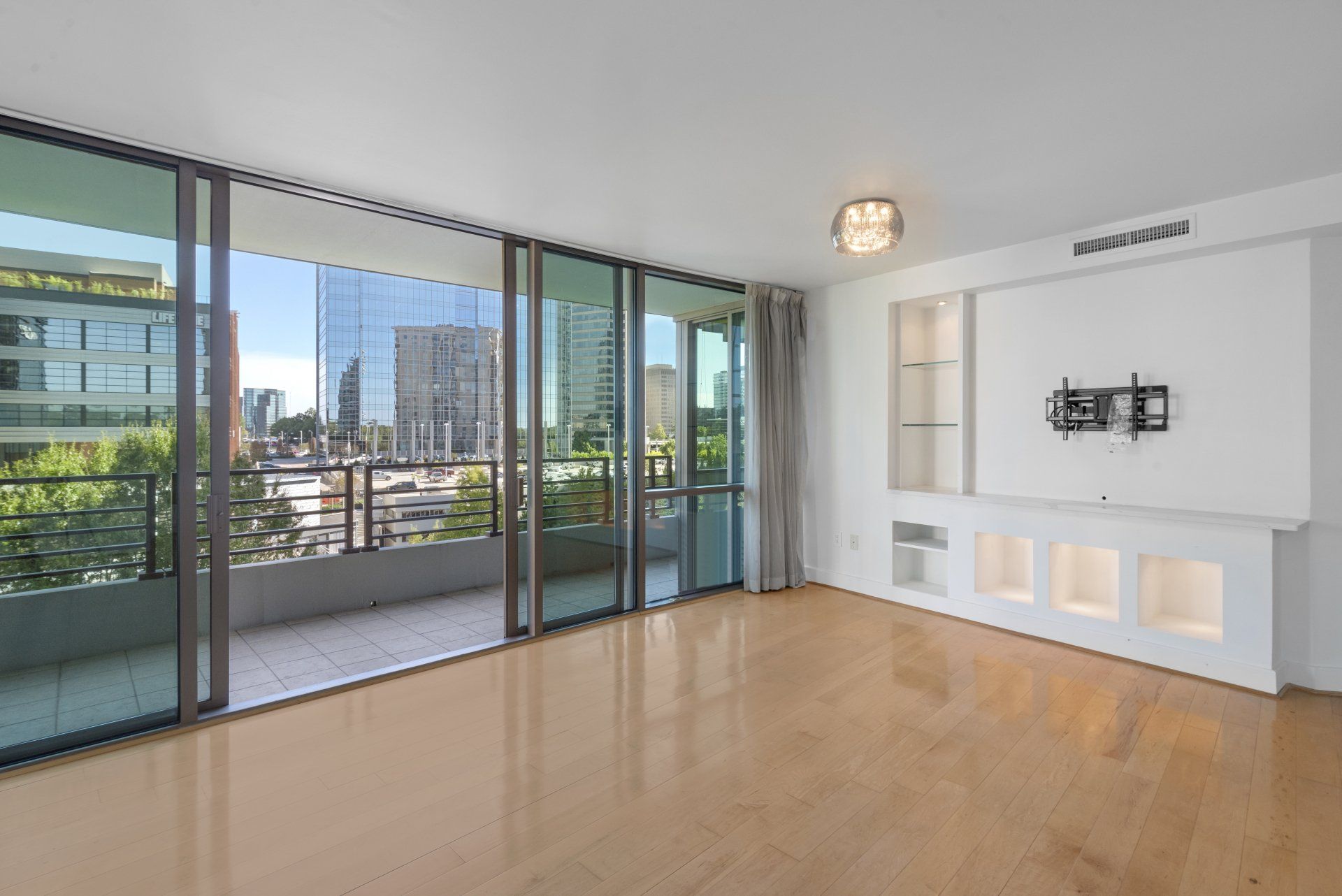 Bright living room with open balcony, city views, light wood floors, and built-in white wall unit.