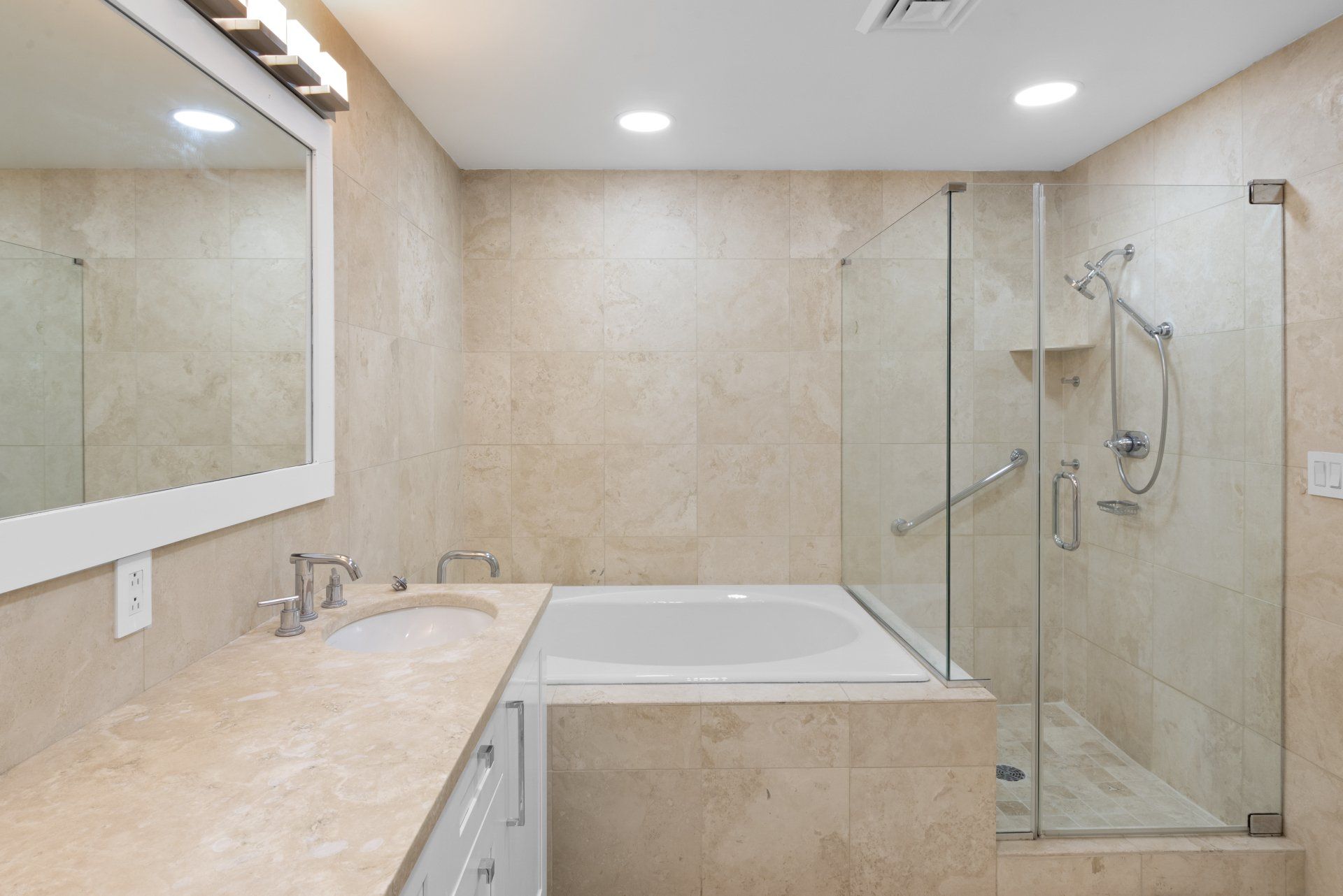 Bathroom with beige tiled walls, a glass shower, a white bathtub, and a long sink counter.