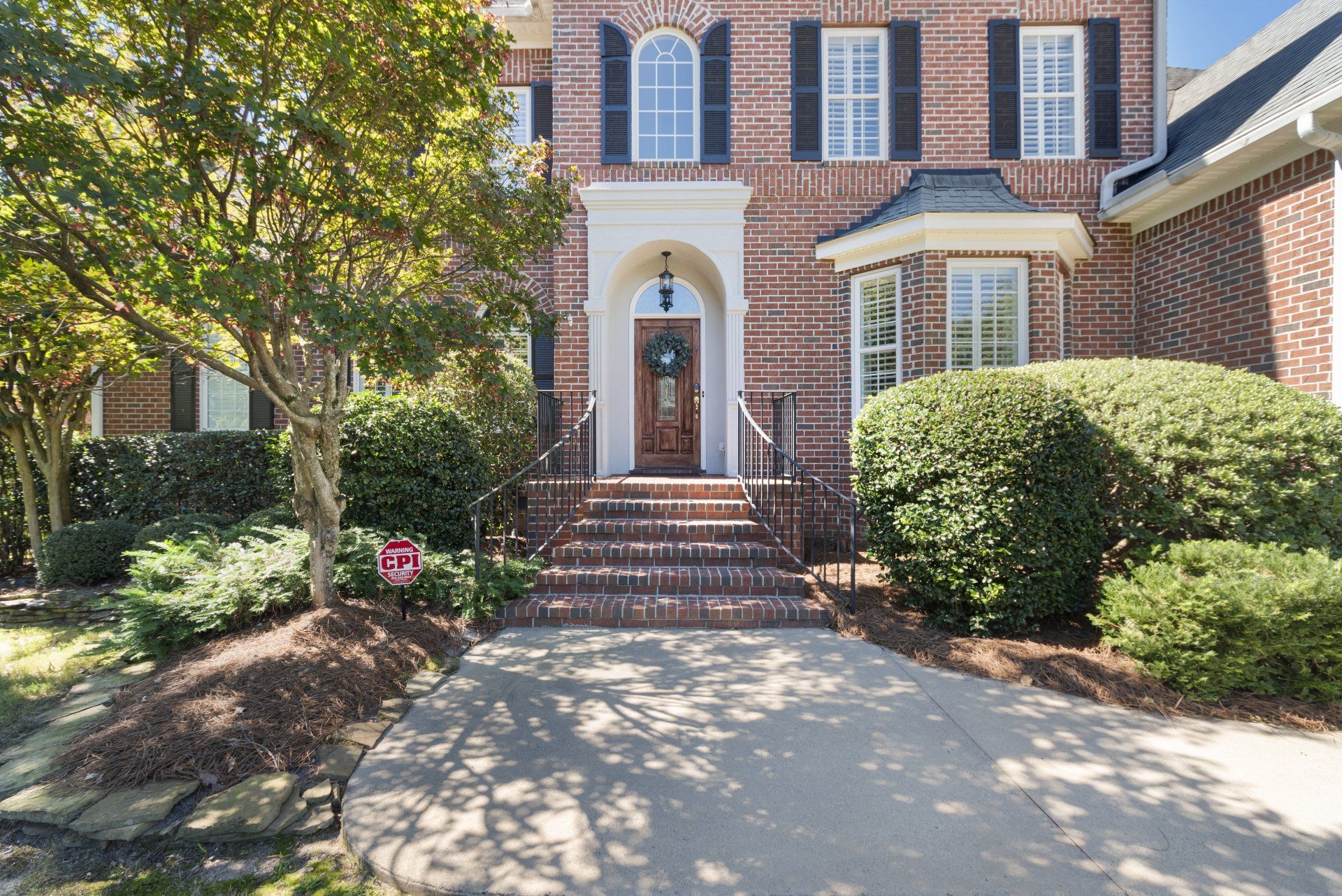 Brick house with steps, arched doorway, and bay window. Green bushes and a tree in front.