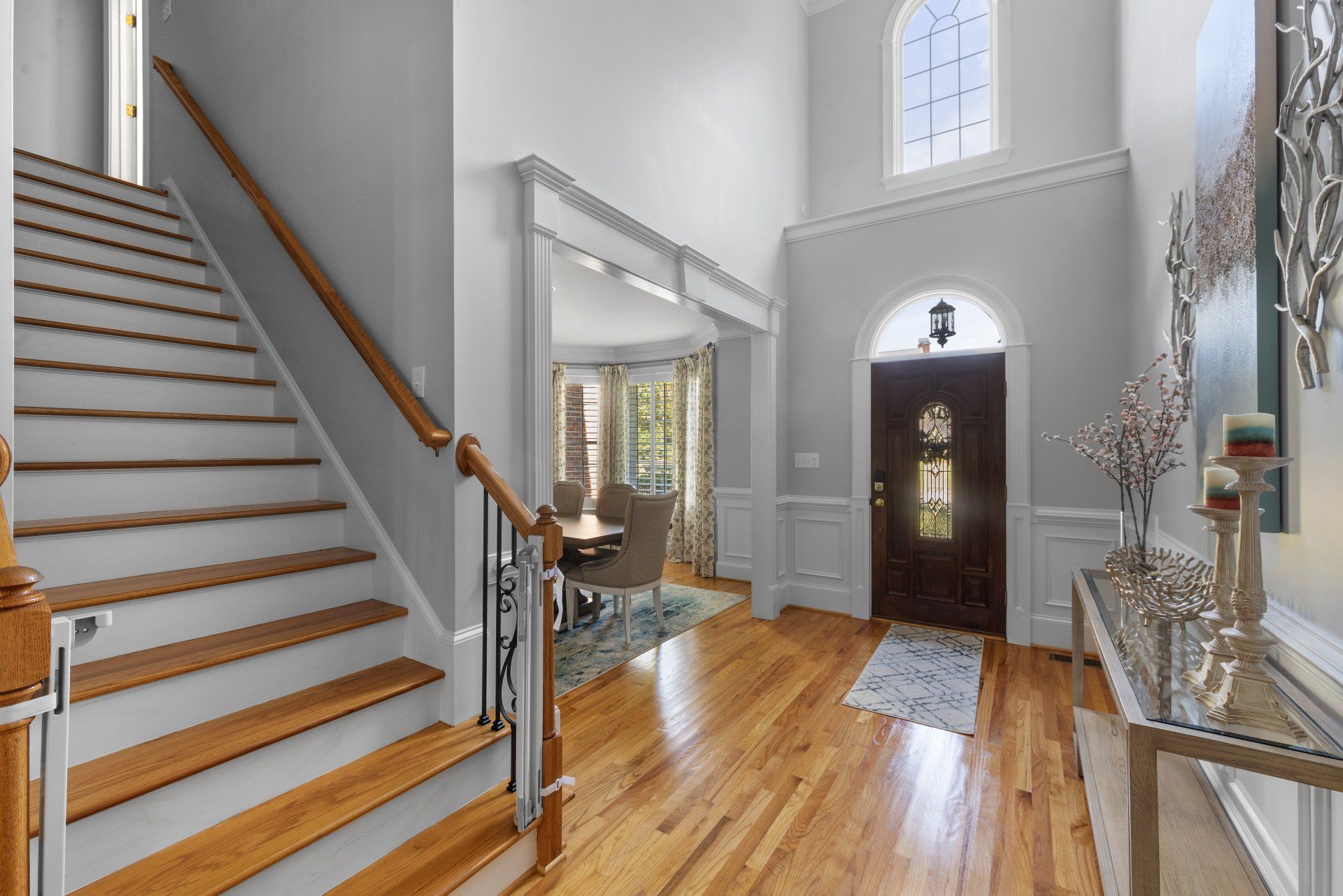 Wooden staircase in a home entryway, leading up to the second floor. Hardwood floors, light gray walls, arched doorway.