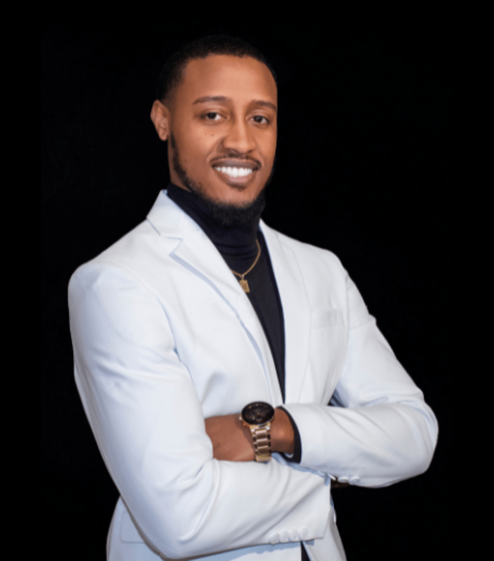 Man in white suit and black turtleneck, arms crossed, smiling, black background.