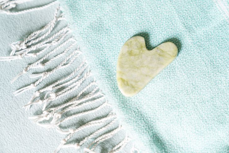 Een hartvormig jade gua sha-instrument op een lichtblauwe handdoek met witte franjes.