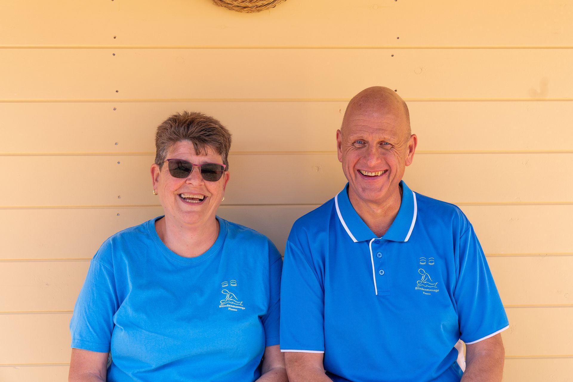 Twee lachende mensen in blauwe shirts staan voor een beige muur.