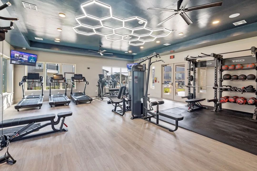A modern apartment fitness center featuring treadmills, weight lifting racks, and geometric honeycomb ceiling lighting.