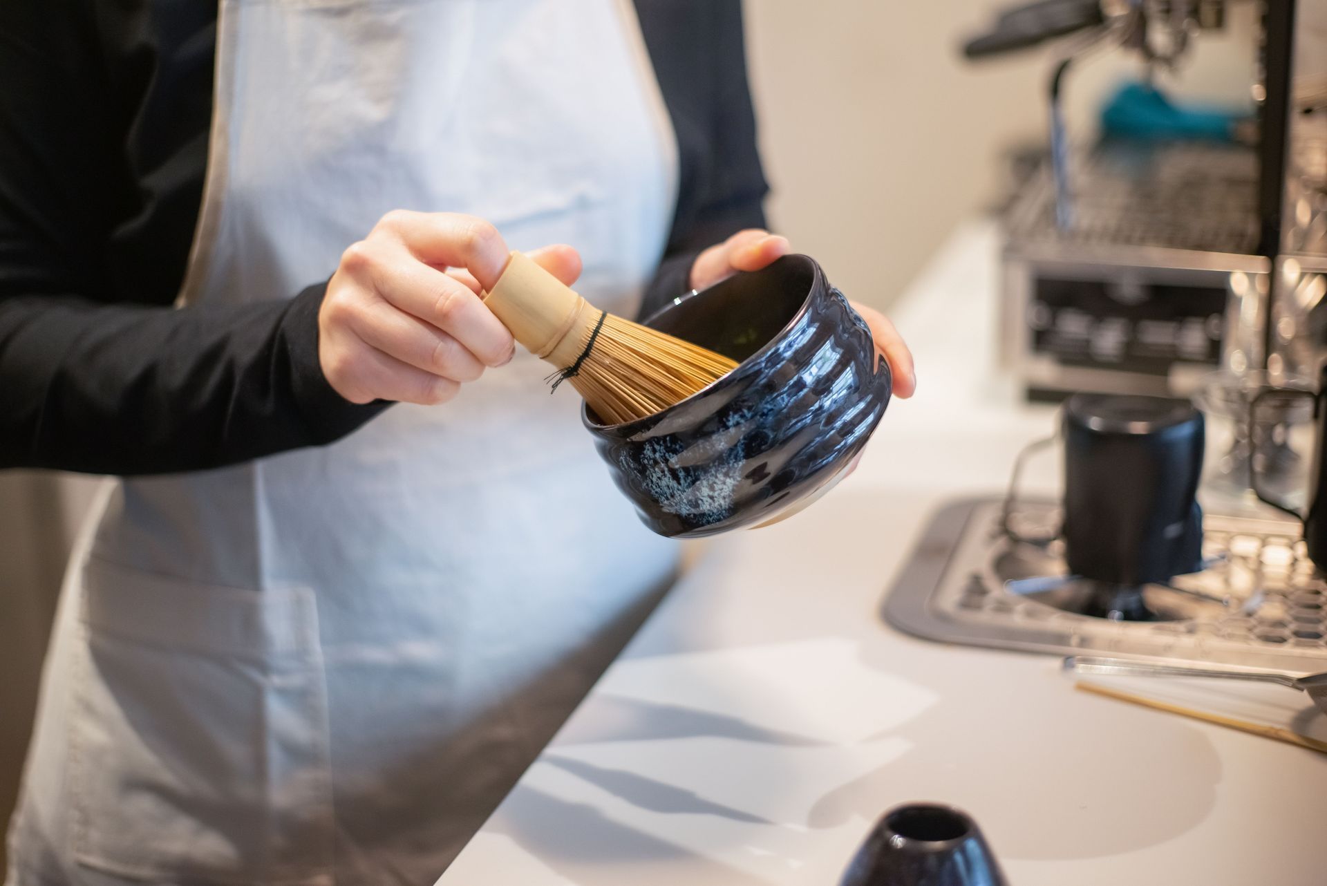 A person wearing a white apron holding a bamboo matcha whisk inside a dark ceramic bowl standing at a kitchen counter.