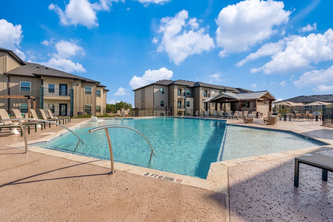 Outdoor apartment pool with clear blue water, tan decking, and lounge chairs under a bright sky