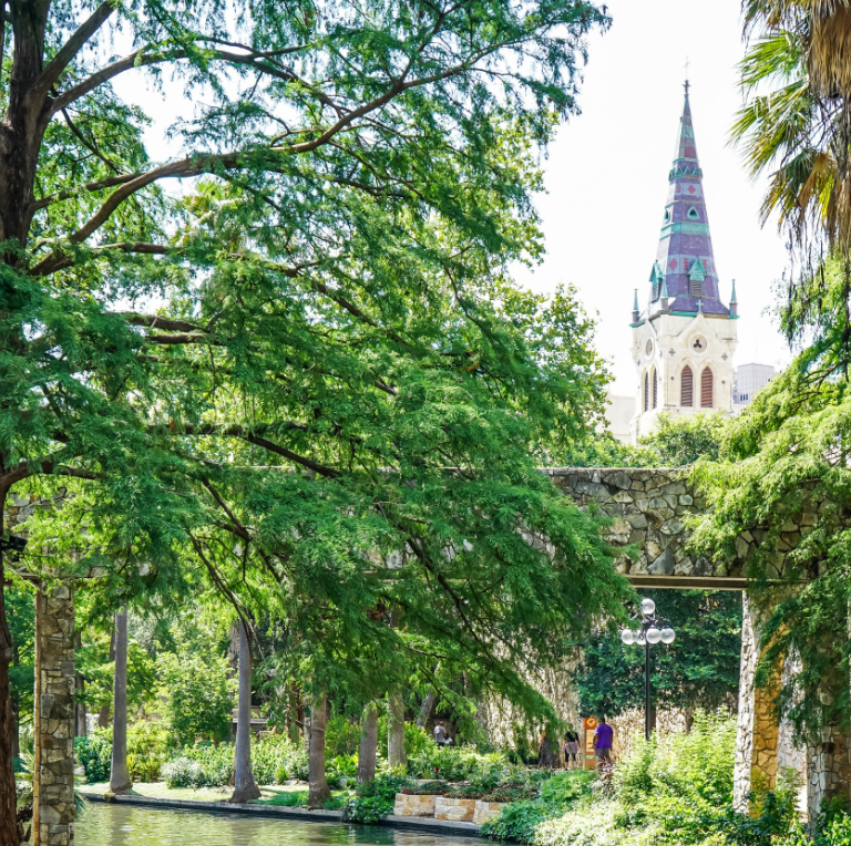Lush park with a stone archway and a colorful church spire rising above the trees.